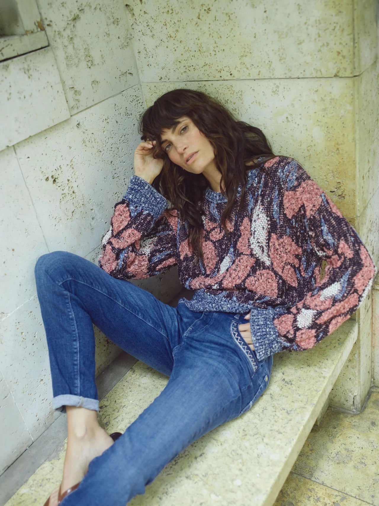 Woman wearing a patterned blouse and blue jeans sitting on a stone bench.