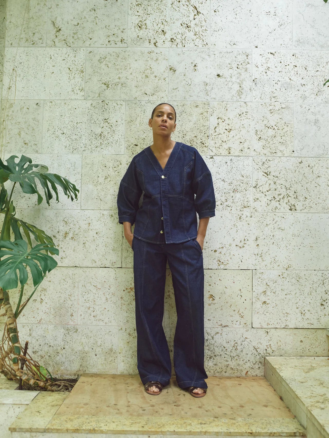 Person wearing a dark denim outfit standing against a textured wall with a plant to the left.