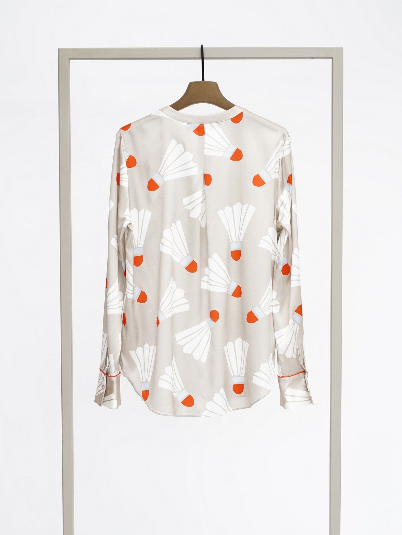 A long-sleeve button-up blouse with a beige base, featuring a white and orange abstract print, displayed on a wooden hanger suspended from a minimalist metal clothing rack against a white studio background.