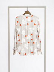 A long-sleeve button-up blouse with a beige base, featuring a white and orange abstract print, displayed on a wooden hanger suspended from a minimalist metal clothing rack against a white studio background.