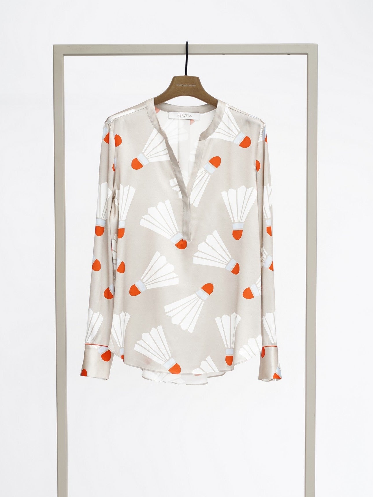 A long-sleeve button-up blouse with a white base and an orange and white abstract pattern, hanging on a wooden hanger from a minimalist gray clothing display stand against a white studio background.
