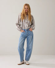 A woman stands against a plain light gray studio background wearing a loose striped blouse, light blue wide-leg jeans, and white sneakers.