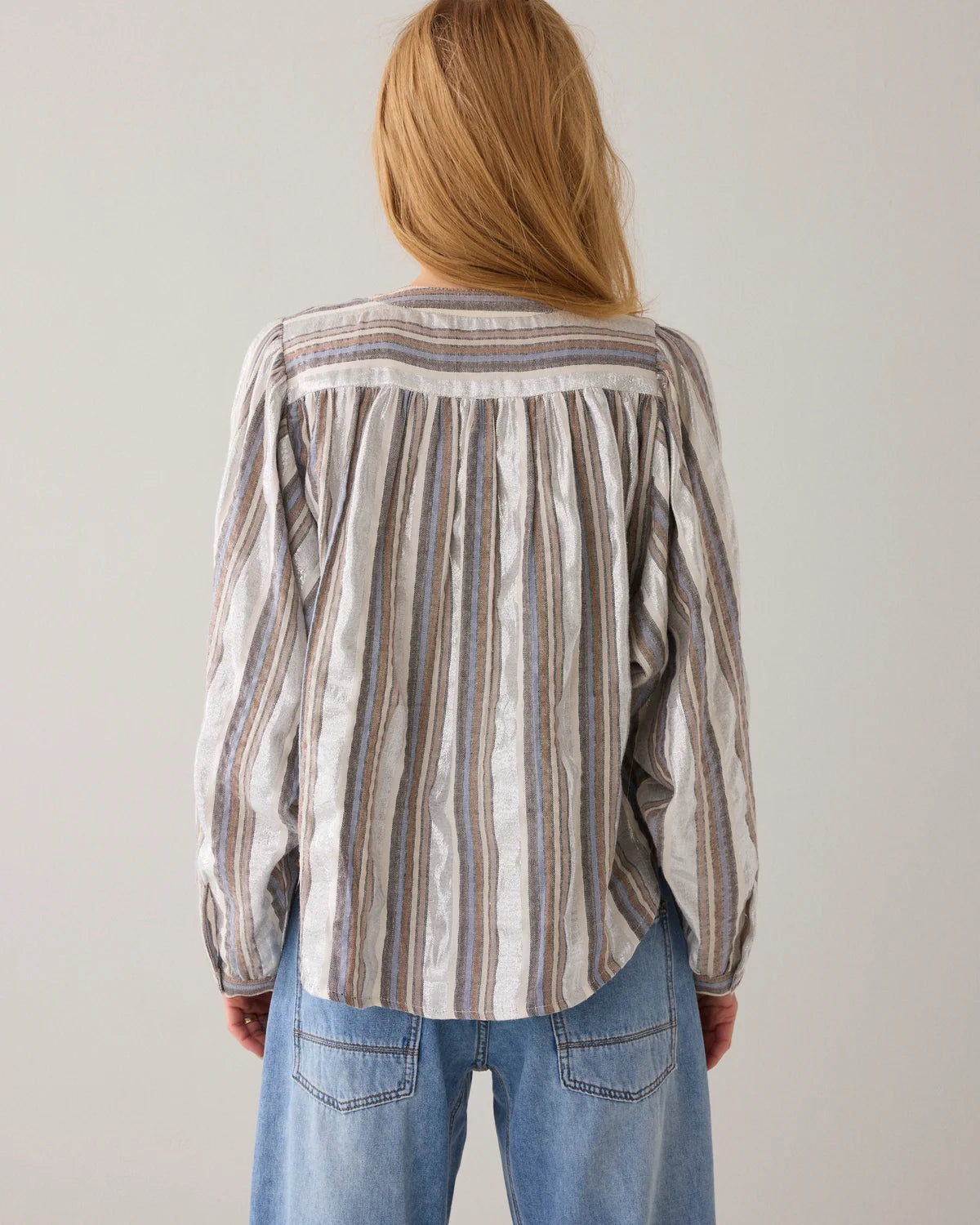 A woman viewed from behind wearing a long-sleeve striped blouse with vertical stripes in beige, taupe, soft blue and silver lurex on a white base, paired with light blue jeans, against a plain light gray background.