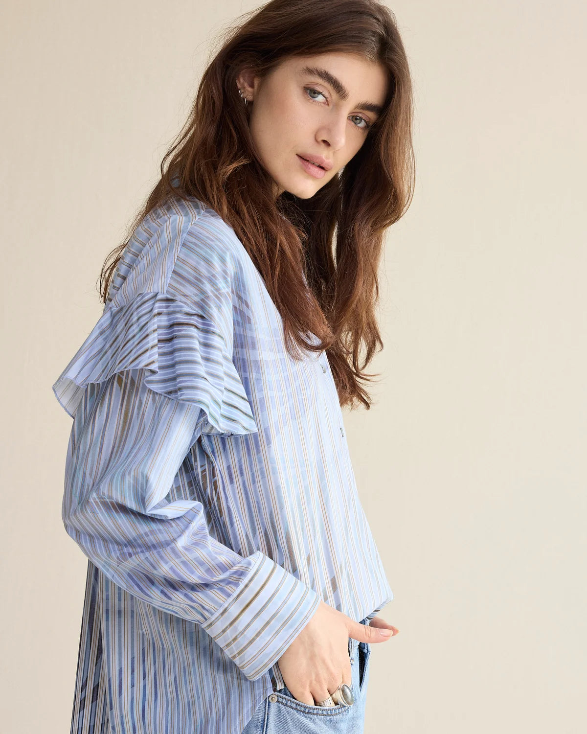 A woman wearing a light blue and white vertically striped long-sleeve blouse with ruffled shoulder details, paired with light-wash jeans, set against a solid beige background.