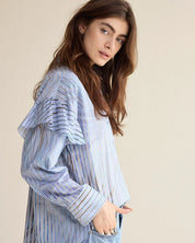 A woman wearing a light blue and white vertically striped long-sleeve blouse with ruffled shoulder details, paired with light-wash jeans, set against a solid beige background.