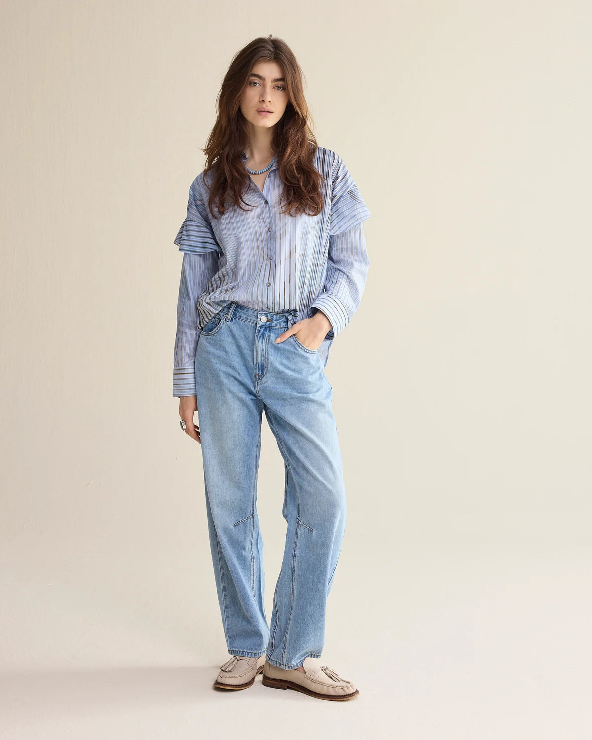 A woman modeling a light blue striped button-up shirt with frill details and light-wash blue jeans, standing against a plain beige studio background.
