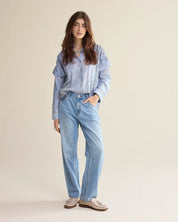 A woman modeling a light blue striped button-up shirt with frill details and light-wash blue jeans, standing against a plain beige studio background.