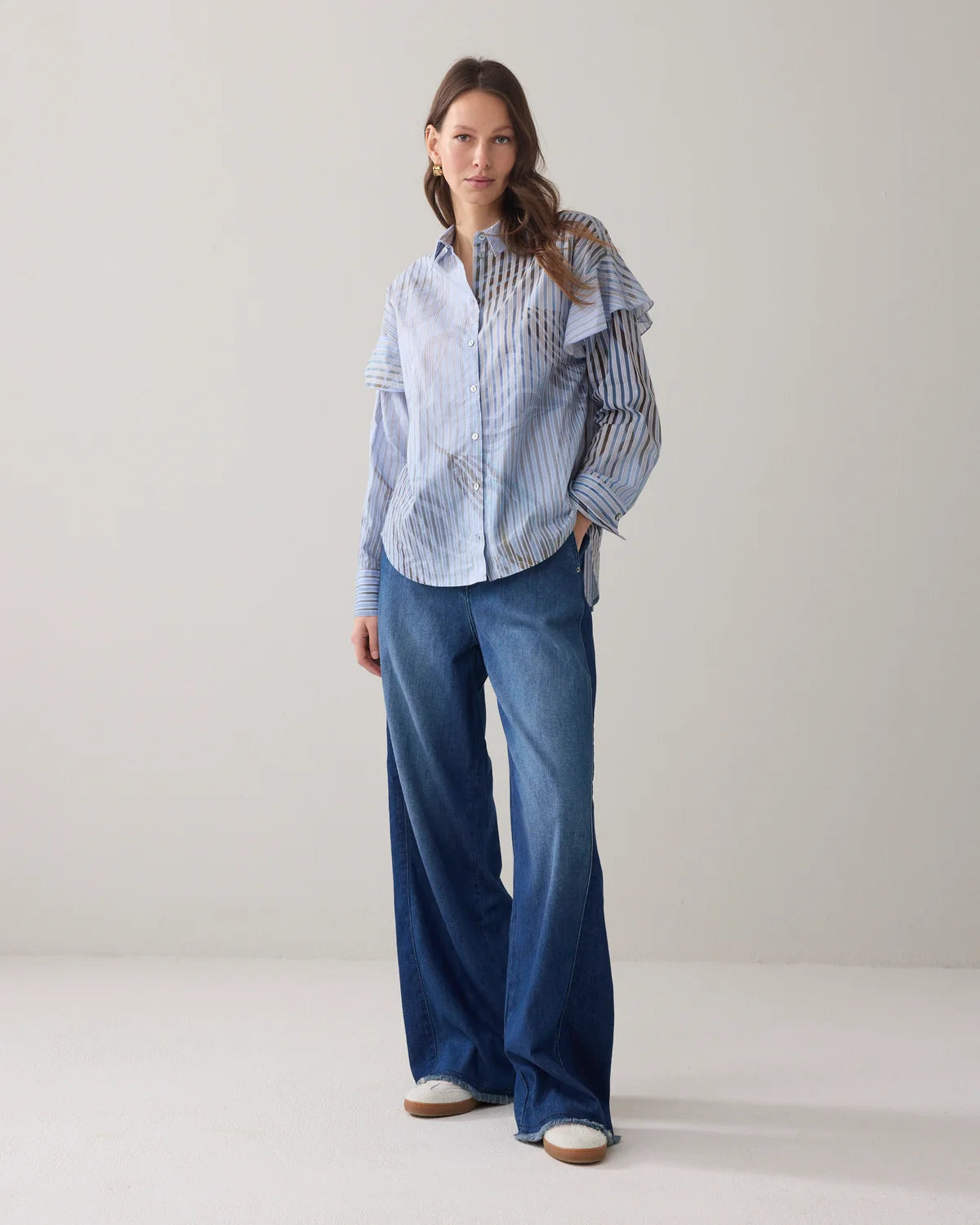 A woman stands in a studio wearing a light blue and white vertically striped button-up shirt with ruffled shoulder details, paired with wide-leg blue jeans and white sneakeres.