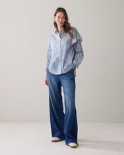 A woman stands in a studio wearing a light blue and white vertically striped button-up shirt with ruffled shoulder details, paired with wide-leg blue jeans and white sneakeres.