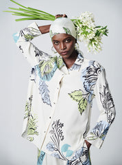 A model wearing a cream-colored button-up shirt and matching headscarf with a blue, green, and black botanical pattern, holding a bouquet of white flowers against a light gray studio background.