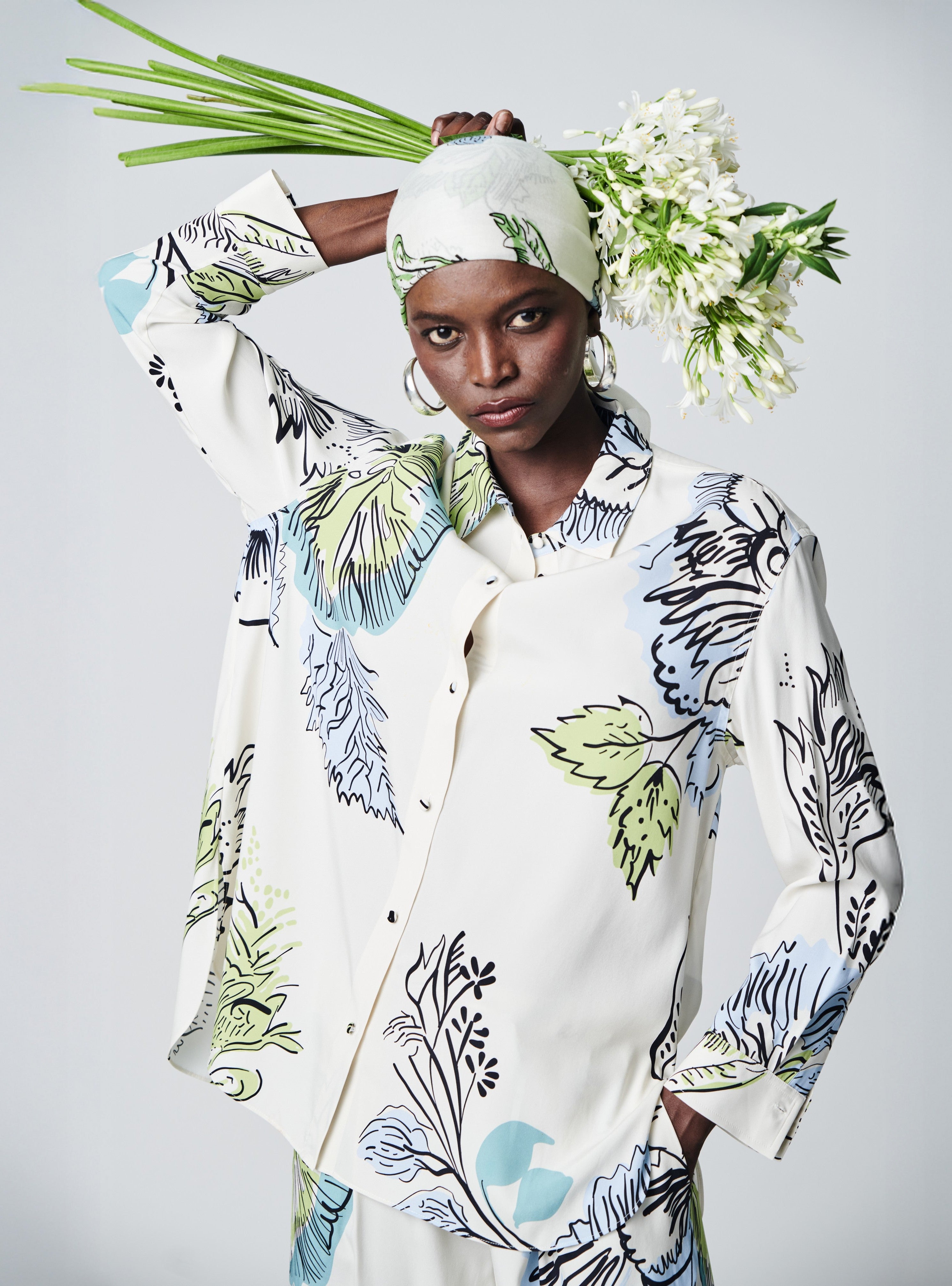 A model wearing a cream-colored button-up shirt and matching headscarf with a blue, green, and black botanical pattern, holding a bouquet of white flowers against a light gray studio background.