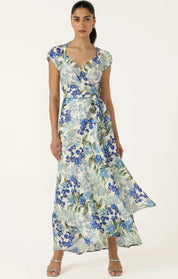 A woman modeling a maxi wrap dress with a blue and green floral print on a light background, paired with silver heeled sandals, against a plain off-white studio background.