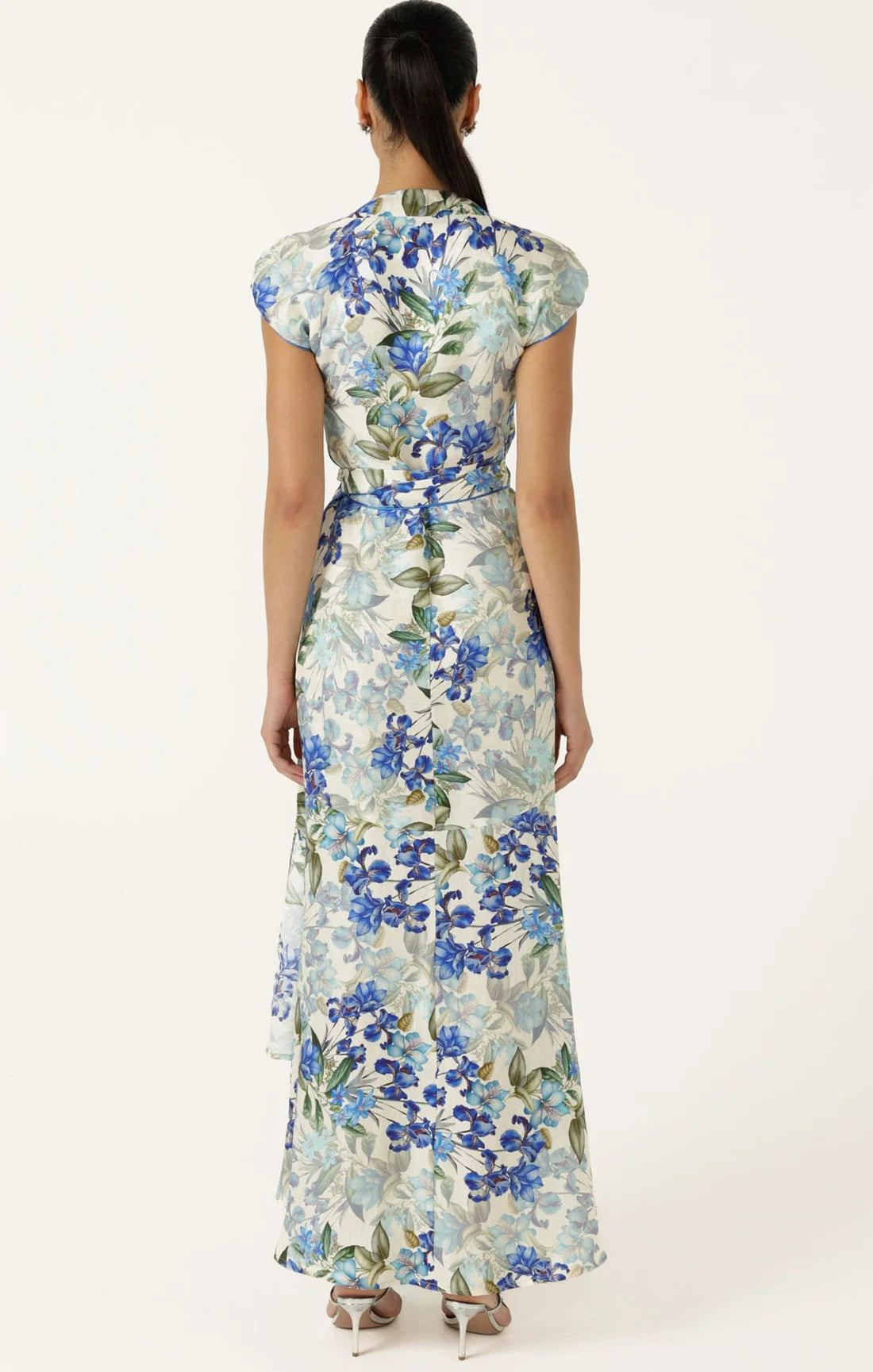 A woman viewed from the back wearing a floor-length floral dress with cap sleeves and a tied waist, on a plain light background.
