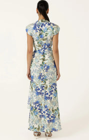 A woman viewed from the back wearing a floor-length floral dress with cap sleeves and a tied waist, on a plain light background.