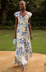 A woman walking outdoors in a flowing cream maxi dress with a blue and green floral print, holding a small light blue clutch.
