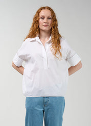 A woman with long red hair stands against a light gray studio background wearing a white short-sleeve button-up shirt, light blue jeans, and white slide sandals.