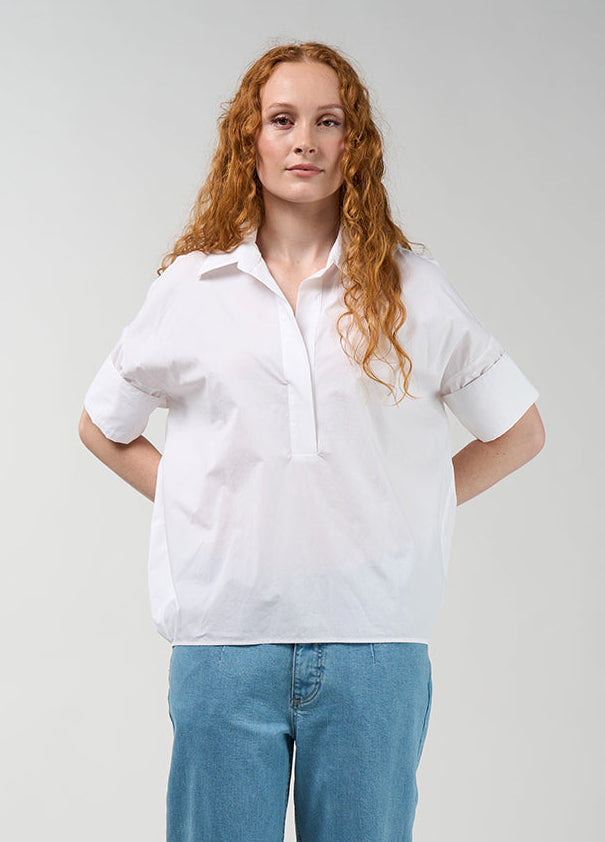 A woman with long red hair stands against a light gray studio background wearing a white short-sleeve button-up shirt, light blue jeans, and white slide sandals.
