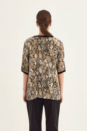 A woman viewed from the back wearing a short-sleeve top with a beige and black snakeskin pattern and black wide-leg pants, against a plain light background.