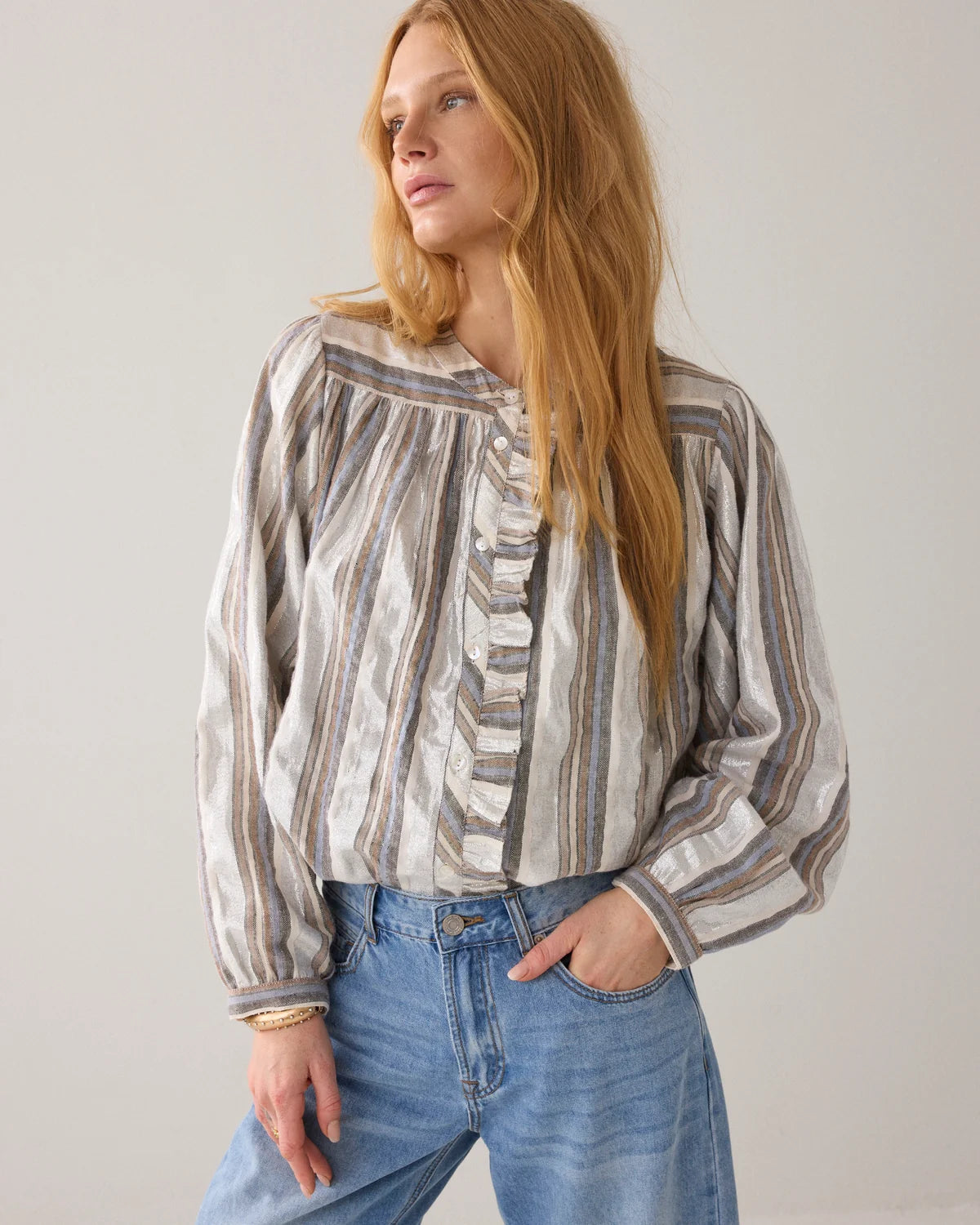A woman with long blonde hair wearing a long-sleeve striped blouse with ruffles and blue jeans, standing against a plain light gray background.