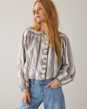 A woman with long blonde hair wearing a long-sleeve striped blouse with ruffles and blue jeans, standing against a plain light gray background.